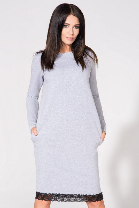 Female model wearing a light grey jersey A-line midi dress with long sleeves and black lace trim at the hem stands against a white background with her hands in pockets