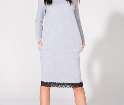 Female model wearing a light grey jersey knit long sleeve midi dress with black lace trim standing with hands in pockets and legs apart against a plain white studio background