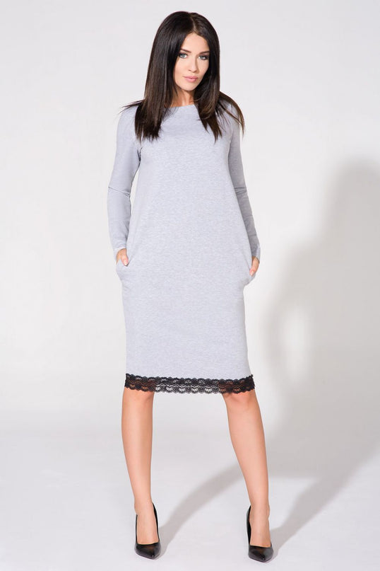 Female model wearing a light grey jersey knit long sleeve midi dress with black lace trim standing with hands in pockets and legs apart against a plain white studio background