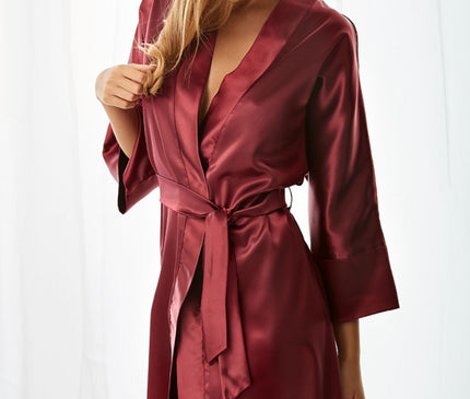 Female model wearing a burgundy satin belted kimono-style robe with three-quarter sleeves and mid-thigh cut gently touching her hair as she stands in front of sheer white curtains
