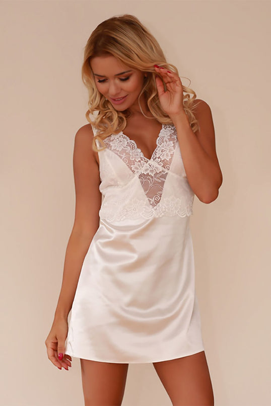A blonde model wears a white satin slip dress with a lace V-neck bodice and short cut standing with one hand to her hair and looking down against a cream wall background