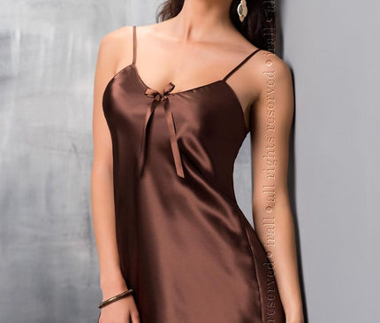 Model in a brown satin mini slip dress with spaghetti straps and bow detail leaning against a grey textured wall