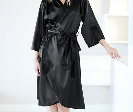 A blonde model stands barefoot in a bright room wearing a black satin wrap robe with three-quarter sleeves and tie belt as she leans on a white countertop with a light wooden floor and a white shelf with a clock in the background