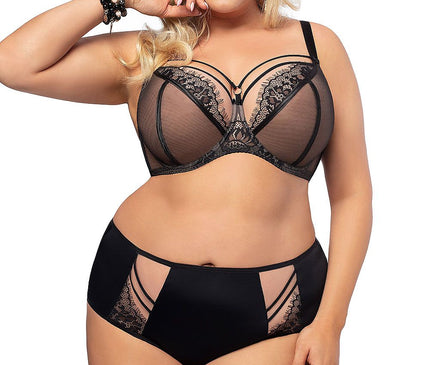 Plus size blonde model standing against a white background wearing a sheer black lace underwired bra with strappy cutout detail and matching high-waisted briefs with mesh panels and lace inserts posing with one hand near her face