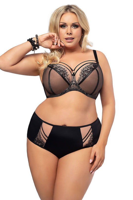 Plus size blonde model standing against a white background wearing a sheer black lace underwired bra with strappy cutout detail and matching high-waisted briefs with mesh panels and lace inserts posing with one hand near her face