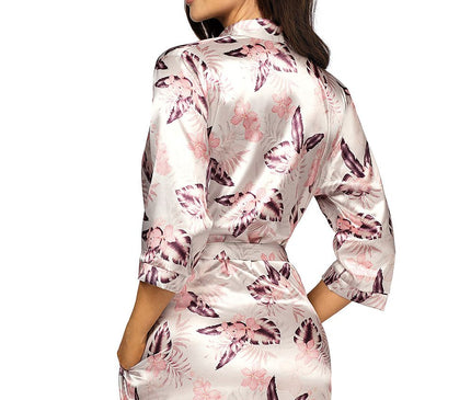 A female model in a pale pink and purple floral satin short kimono robe tied at the waist stands against a white background with her back to camera looking over her shoulder