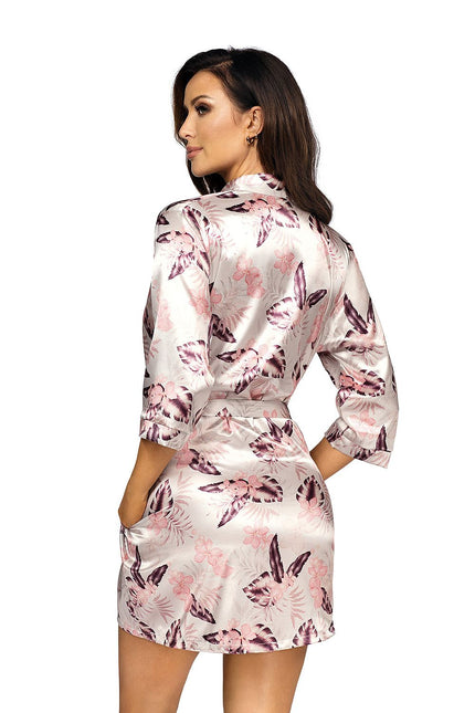 A female model in a pale pink and purple floral satin short kimono robe tied at the waist stands against a white background with her back to camera looking over her shoulder