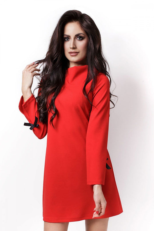 Female model standing against a white background wearing a red long-sleeve jersey A-line shift dress with black bow details