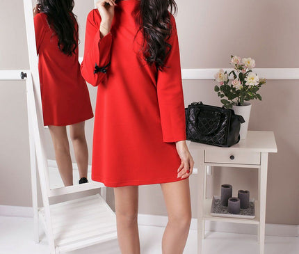 A model wearing a red long-sleeved shift dress in smooth fabric poses beside a white full-length mirror and a small white table with flowers and a black handbag against a pale grey wall