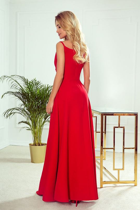 Blonde model in a red sleeveless crepe A-line floor-length gown stands with her back to camera and head turned over her shoulder against a white paneled wall beside a gold framed glass table and potted palm plant