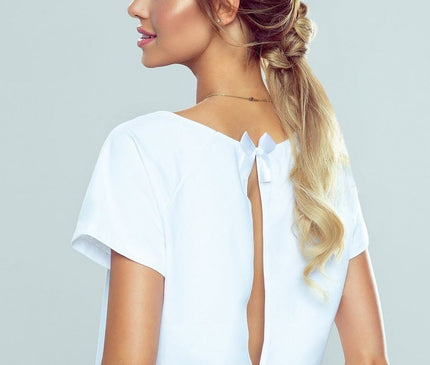 A female model in a cotton short-sleeve white top with a V-back cut and bow detail paired with jeans stands in profile against a plain grey background