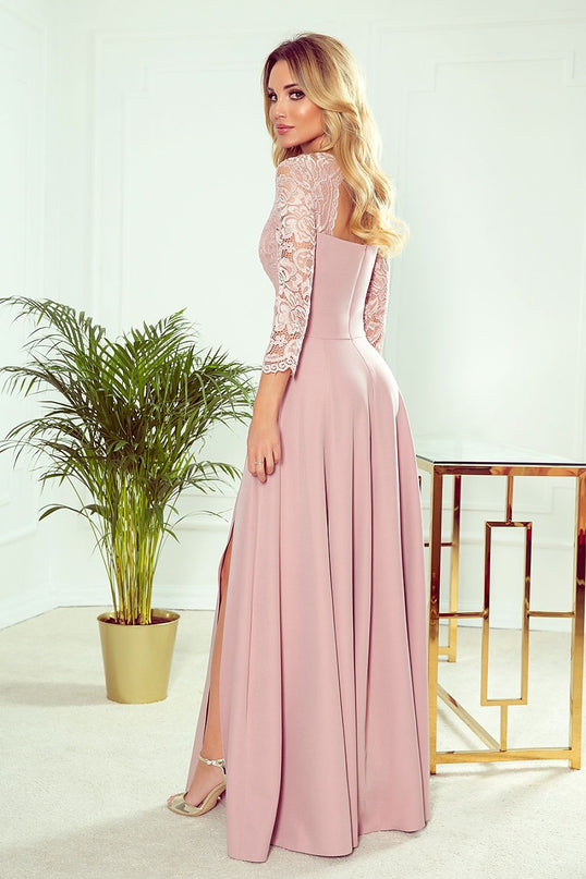 Blonde model wearing a pale pink maxi dress with lace bodice, fitted waist and flowing skirt with a side slit standing in a white room with a potted palm and a gold metal table