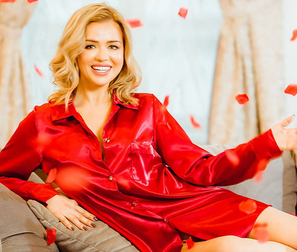 A female model reclines on a grey sofa in a red satin button-up shirt dress with a relaxed cut leaning against cushions in front of beige curtains with red rose petals scattered around