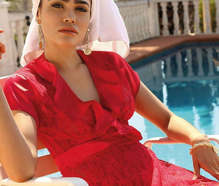 A model sits on a white plastic chair by a pool wearing a red lace short-sleeve dress with a ruffled collar and a white towel turban against a backdrop of palm trees and a white balustrade