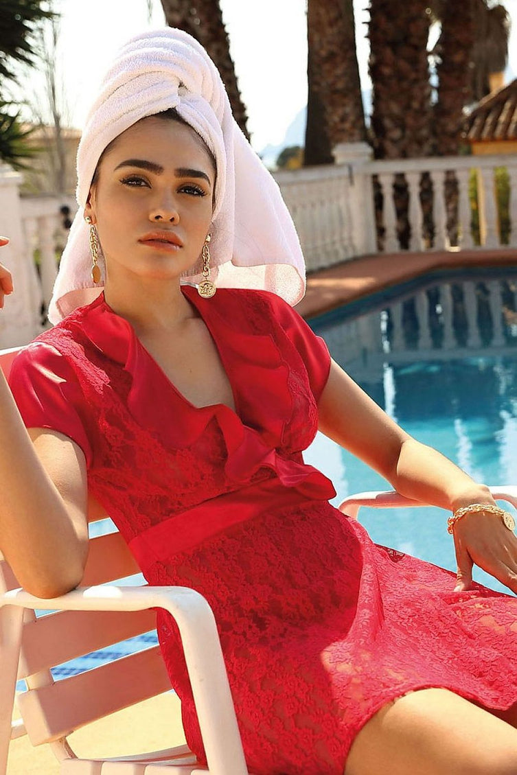 A model sits on a white plastic chair by a pool wearing a red lace short-sleeve dress with a ruffled collar and a white towel turban against a backdrop of palm trees and a white balustrade