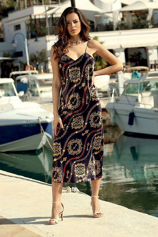 A female model in a black satin slip maxi dress with gold and red chain print and spaghetti straps stands with one hand on her hip on a sunlit marina dock with moored yachts and shimmering water in the background