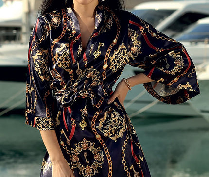 Model stands with one hand on her hip wearing a navy satin mini kimono robe with gold chain print beside a marina with yachts and water