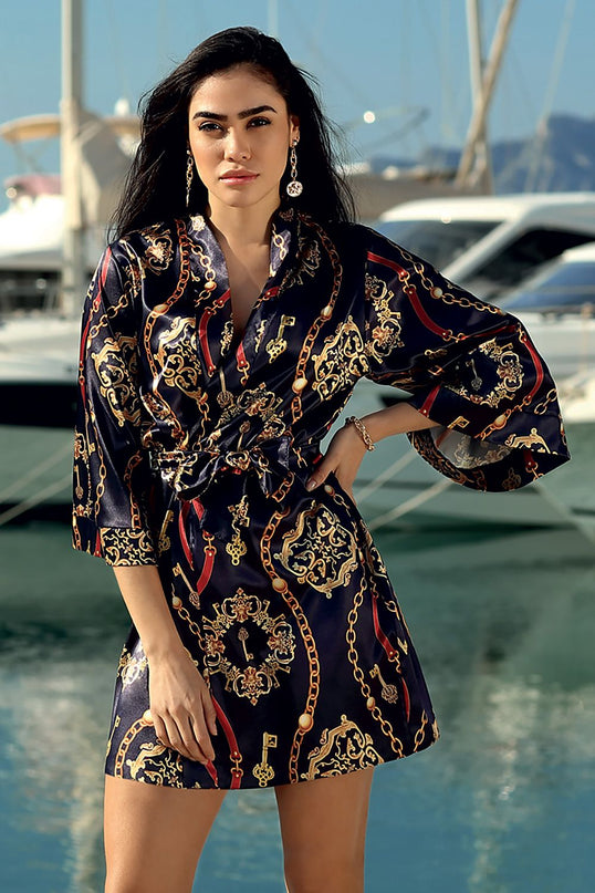 Model stands with one hand on her hip wearing a navy satin mini kimono robe with gold chain print beside a marina with yachts and water