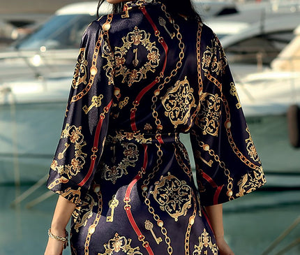 A female model turned back to camera wearing a navy satin kimono-style mini dress with gold chain and key print standing by a marina with moored yachts and water