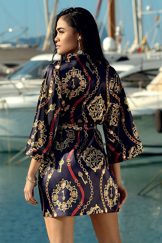 A female model turned back to camera wearing a navy satin kimono-style mini dress with gold chain and key print standing by a marina with moored yachts and water