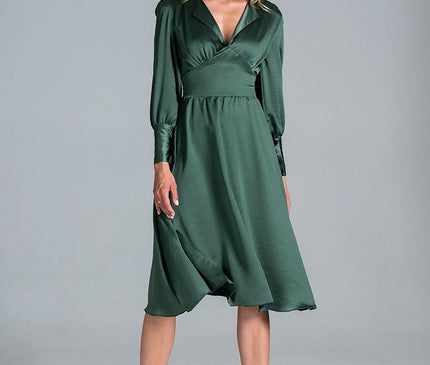 A blonde model stands centred against a plain light grey studio backdrop wearing a dark green satin long sleeve V-neck midi dress with a fitted waist