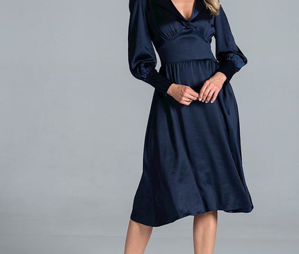 A female model stands in a plain light grey studio wearing a navy blue satin midi dress with an empire waist and V-neckline, lightly clasping her hands in front