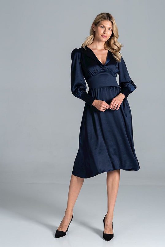 A female model stands in a plain light grey studio wearing a navy blue satin midi dress with an empire waist and V-neckline, lightly clasping her hands in front