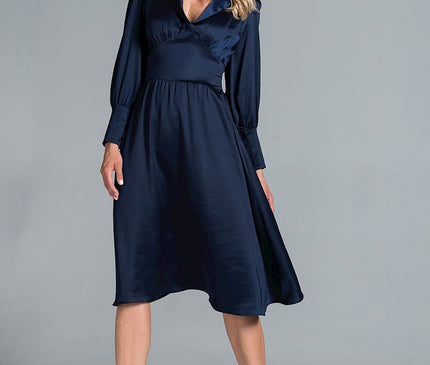 Model stands in a studio setting wearing a navy blue satin A-line midi dress with long sleeves and a fitted waist looking down against a grey background