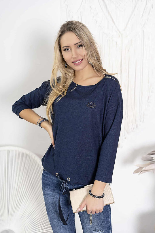 Model wearing a navy blue jersey boat-neck top with three-quarter sleeves and drawstring hem standing against a white wall with macrame hanging and a white fan-back chair