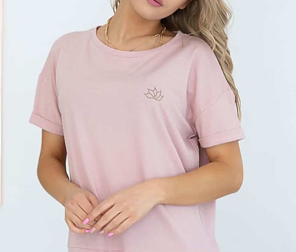 A blonde female model wears a relaxed-fit pink cotton short-sleeve top with a drawstring hem while standing with clasped hands and a tilted head against a white wall and wire chair