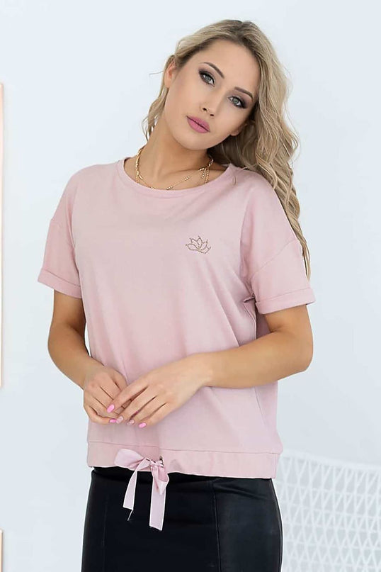 A blonde female model wears a relaxed-fit pink cotton short-sleeve top with a drawstring hem while standing with clasped hands and a tilted head against a white wall and wire chair