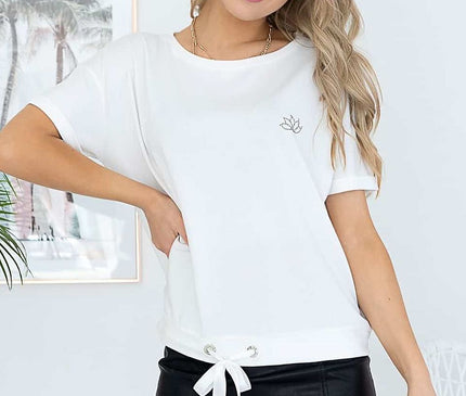 A blonde female model standing with one hand on hip and head tilted wears a loose cut white cotton T-shirt with drawstring hem and small embroidered motif paired with a black faux leather mini skirt in a white room with framed palm tree prints and a leafy plant