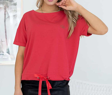 A blonde model stands against a white wall and wicker chair wearing a red jersey drawstring-hem T-shirt and a black leather mini skirt with one hand touching her hair
