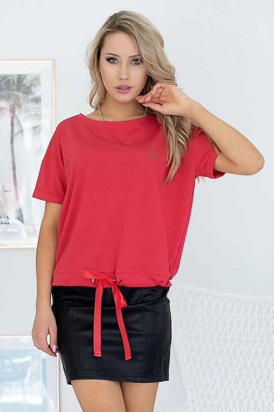 A blonde model stands against a white wall and wicker chair wearing a red jersey drawstring-hem T-shirt and a black leather mini skirt with one hand touching her hair