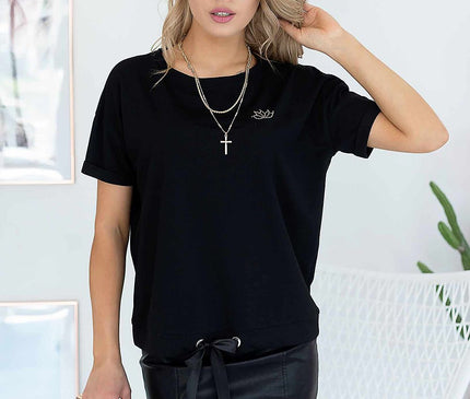 Blonde female model stands facing camera in a white room wearing a black cotton jersey T-shirt with drawstring hem and a fitted black faux leather mini skirt beside a white wicker chair framed wall art and a potted plant