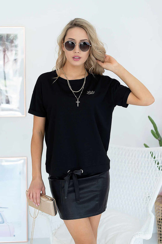 Blonde female model stands facing camera in a white room wearing a black cotton jersey T-shirt with drawstring hem and a fitted black faux leather mini skirt beside a white wicker chair framed wall art and a potted plant