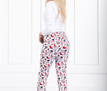 A blonde model stands barefoot in front of a white panelled wall wearing straight-leg white cotton-blend lounge trousers with red and black winter-themed print and looks over her shoulder