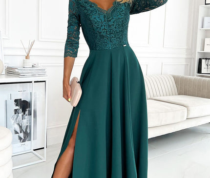 Blonde model stands with one hand in her hair wearing an emerald green lace top and flowing chiffon floor-length evening gown with three-quarter sleeves and a front slit in a white paneled room with a cream sofa, side table with books and reed diffuser and marble floor