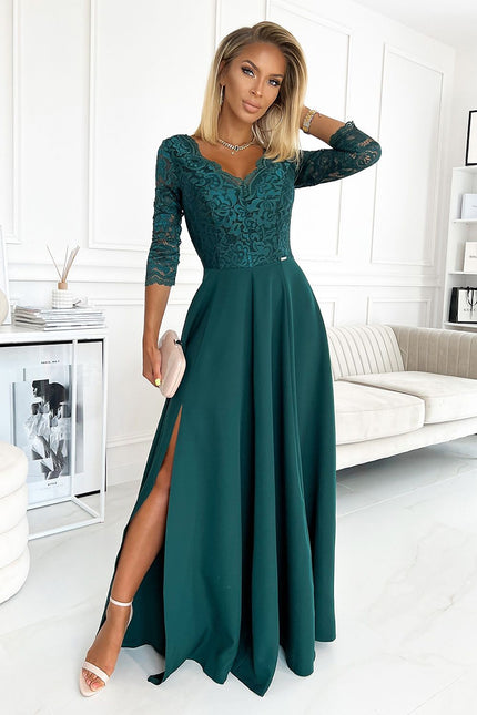 Blonde model stands with one hand in her hair wearing an emerald green lace top and flowing chiffon floor-length evening gown with three-quarter sleeves and a front slit in a white paneled room with a cream sofa, side table with books and reed diffuser and marble floor