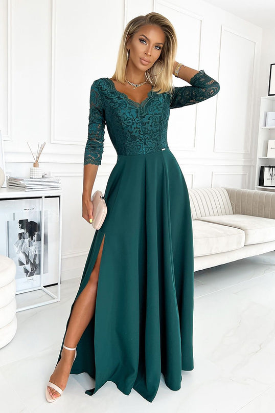 Blonde model stands with one hand in her hair wearing an emerald green lace top and flowing chiffon floor-length evening gown with three-quarter sleeves and a front slit in a white paneled room with a cream sofa, side table with books and reed diffuser and marble floor