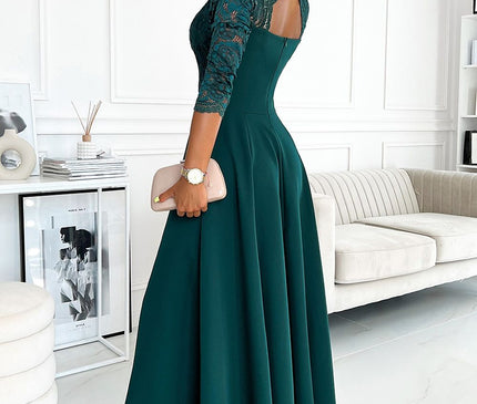 A blonde model wearing a fitted dark green evening gown with lace bodice and flared maxi skirt standing turned over her shoulder in a white room with a sofa and framed artwork