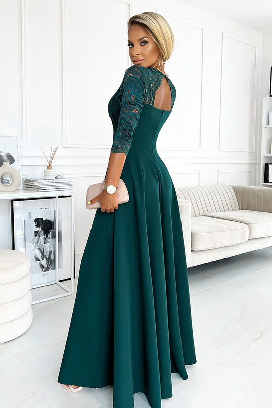 A blonde model wearing a fitted dark green evening gown with lace bodice and flared maxi skirt standing turned over her shoulder in a white room with a sofa and framed artwork