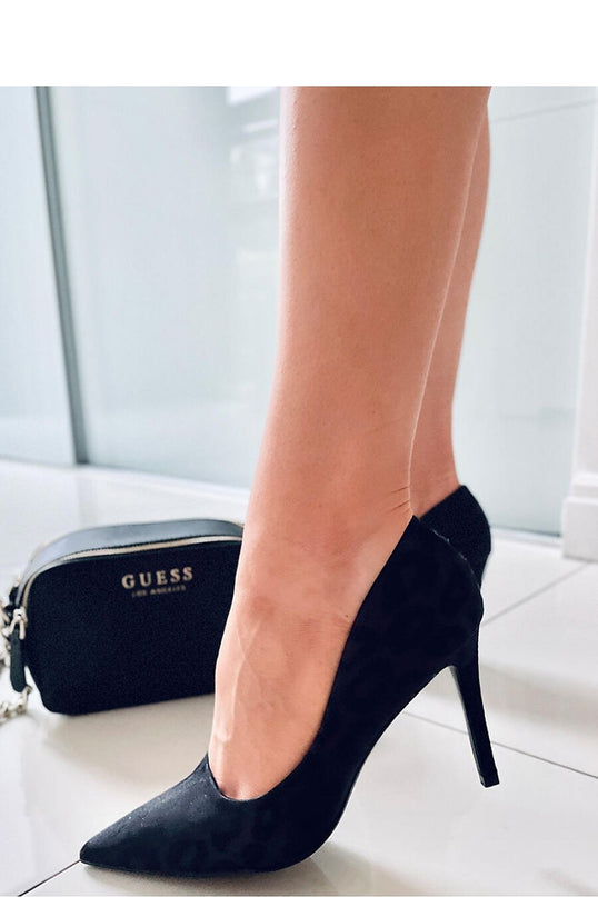 A model stands in black suede pointed-toe stiletto pumps with low-cut sides on a white tiled floor with a small black bag and glass-panelled wall in the background