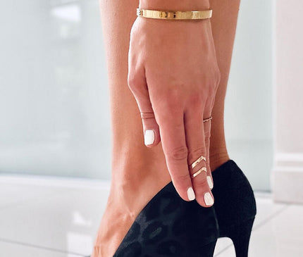 Model wearing black suede leopard-print pointed-toe stiletto pumps adjusting the shoe with a hand adorned with gold rings and white nail polish against a white tiled floor and bright window wall