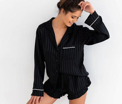 Model posing against a white wall in a black striped satin button-down long-sleeve top and matching ruffled-hem shorts