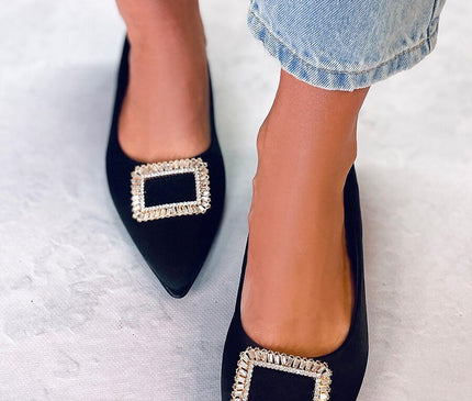 Female model wearing black velvet pointed-toe flats with rectangular crystal buckles stands on a pale textured floor beneath light denim cropped jeans