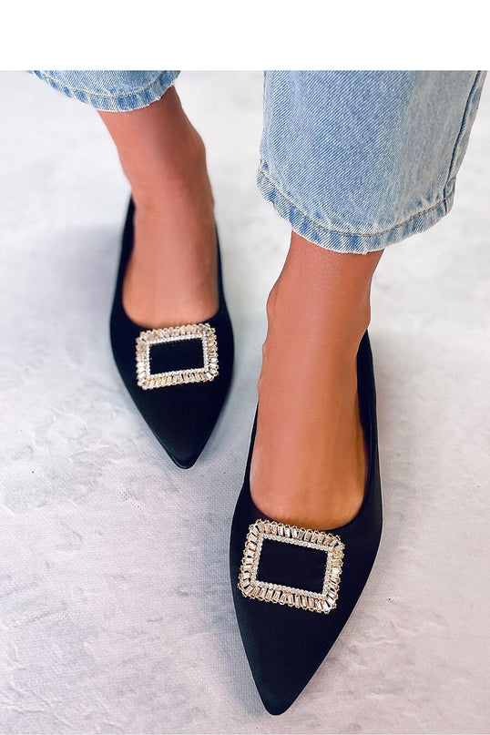 Female model wearing black velvet pointed-toe flats with rectangular crystal buckles stands on a pale textured floor beneath light denim cropped jeans
