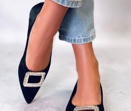 Model wearing black suede pointed toe flats with rectangular crystal buckles poses with crossed ankles on a light grey floor against a white backdrop