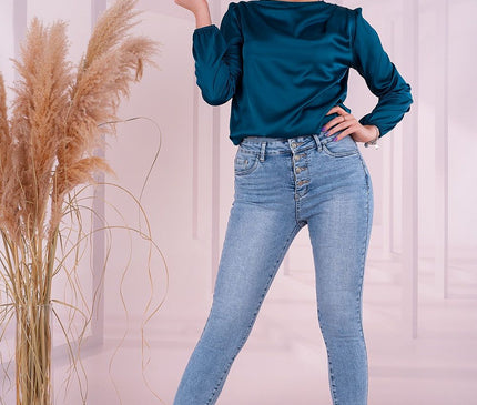 Female model standing with one hand on hip and the other raised wearing a teal satin long-sleeve blouse and light blue high-waisted skinny jeans with black heels against a pale pink room backdrop with a glass vase of dried pampas grass
