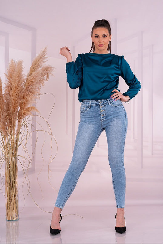 Female model standing with one hand on hip and the other raised wearing a teal satin long-sleeve blouse and light blue high-waisted skinny jeans with black heels against a pale pink room backdrop with a glass vase of dried pampas grass
